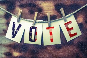 The word VOTE stamped on card stock hanging from old twine and clothes pins over a rusty vintage background.