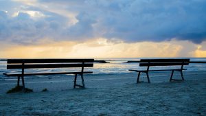 Image is of two benches on the beach facing the ocean.