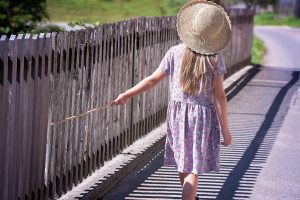 Image is of a a young girl in a sundress and sun hat walking away from the camera holding a stick in her hand that she is running against a wooden fence
