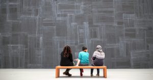 Image is of three women sitting on a bench with their back to the camera, facing a giant art exhibit.