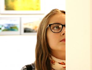 Image is of a girl looking at a piece of art, facing the camera, with art out of focus in the background.