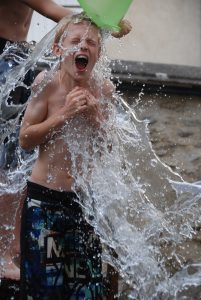Image is of a boy in swim trunks being doused with a bucket of cold water.