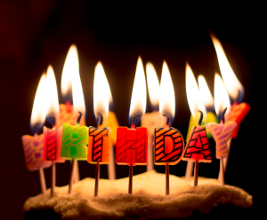 Image is of a lit candles on the cate that spell birthday in front of a black background.