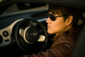 Image is of a male driver with sunglasses sitting behind the wheel of a car.