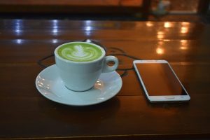 Image is of a green tea latte on a wooden table next to a cell phone.