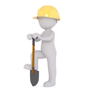 Image is a person in a yellow hardhat breaking ground with a shovel.
