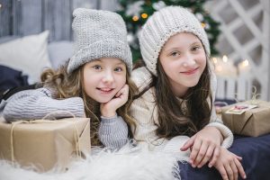 Image is of two girls in winter hats next to presents wrapped in brown paper with Christmas tree in background.