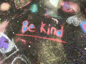 Image is a chalk drawing on asphalt that says Be Kind.
