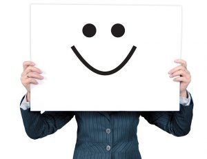 Image is a woman holding up a white board with a black smiley face on it in front of her head.