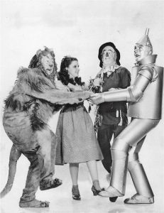 Image is a black and white photo of Dorothy, the Scarecrow, the Tin man, and the Cowardly Lion from the Wizard of Oz