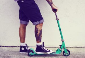 Image is a man in shorts with a leg tattoo standing on a blue-green scooter.