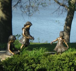 Image is a bronze sculpture of three young girls playing next to a lake.