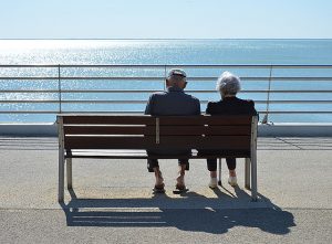 Image is a senior couple sitting on a bench overlooking the ocean on a sunny day.