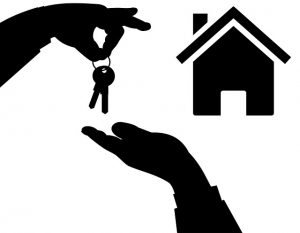 Image is a black and white image of a hand giving house keys to another hand with a home in the background.