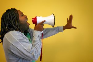 Image is a Black man with dreadlocks yelling into a megaphone against a yellow background.