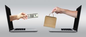 Image is of two computers facing each other, one with a hand holding money coming out of it and the other with a hand holding a shopping back coming out, as a visual representation of online shopping.