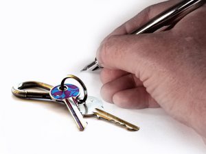 Image is a hand holding a pen getting ready to sign next to some house keys.