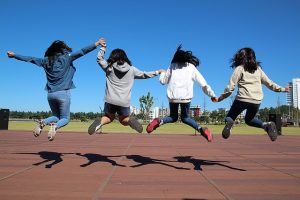 Image is four girls jumping in unison while holding hands with backs to the camera.