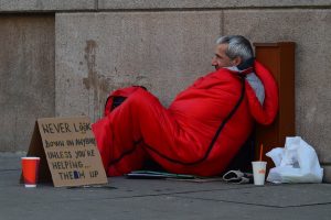 Image is a homeless man on a sidewalk in a sleeping bag with a sign and cup next to him.