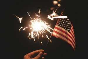 Image is a close up of a person's hand holding a sparkler and a small United States Flag.