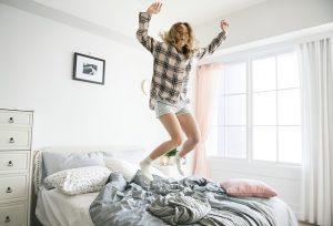 Image is a college student jumping on a bed in an apartment.