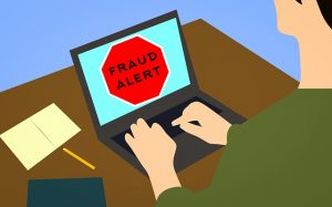 Image is an illustration of someone on a computer with a fraud alert symbol on the screen.