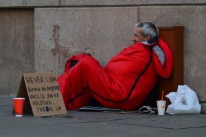 Image is a homeless man sitting in a red sleeping bad on a street with a cardboard sign.
