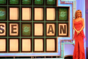 Image is Vanna White next to the puzzle board for Wheel of Fortune.